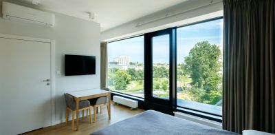 Studio z łóżkiem, telewizorem i stolikiem przy przeszklonej ścianie z widokiem na park